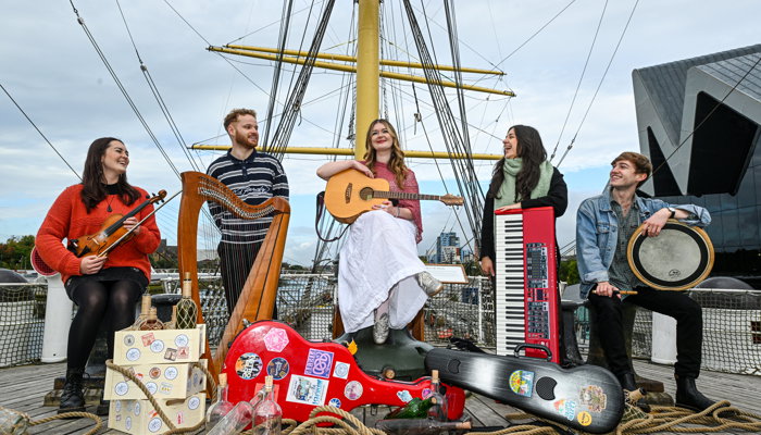 Five musicians performing on the deck of a tall ship surrounded by ropes, instrument cases and maritime rigging.