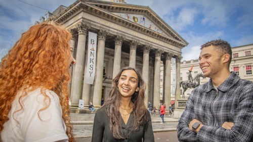 Three people smiling while standing in front of the Gallery of Modern Art in Glasgow on a sunny day.