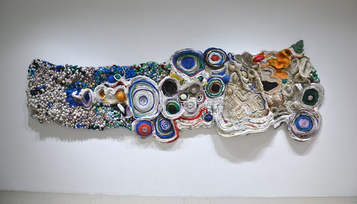 A brightly coloured woven sculpture hangs on a gallery wall