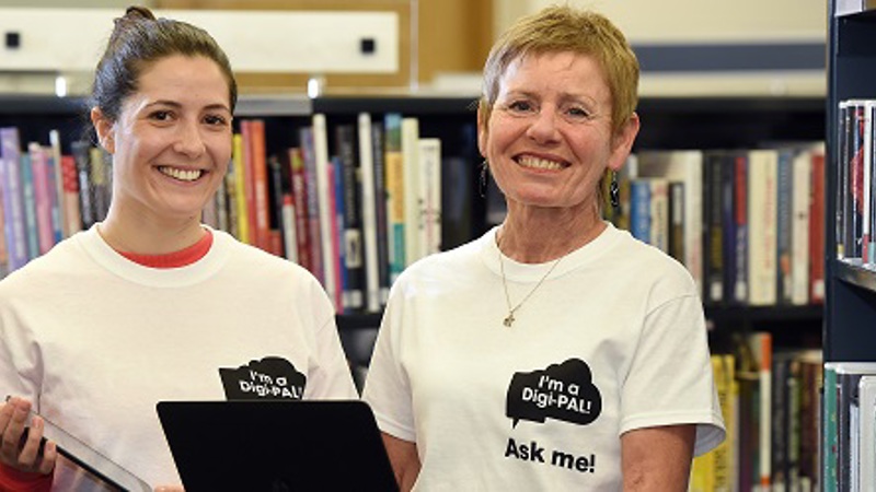 Two Digi-PALS volunteers wearing white t-shirts stand in front of bookshelves in a library.