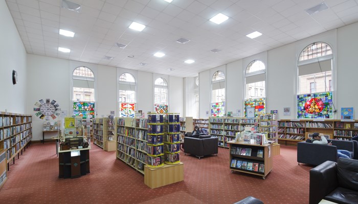 A room in a library with multiple bookcases, red carpet and tall windows. The walls are white.