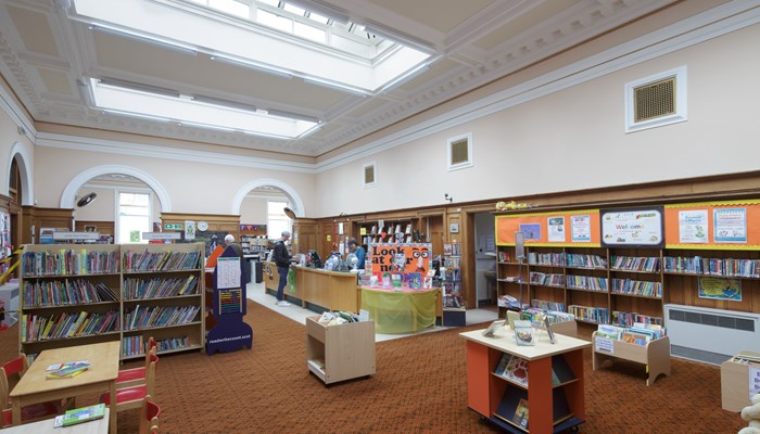 An interior shot of Pollokshields Library with bookcases and tables. In the background, there is a person standing at the information desk.