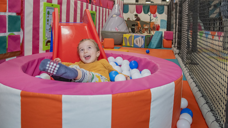 A young child smiling after sliding down a chute into a small ball pit at soft play area.