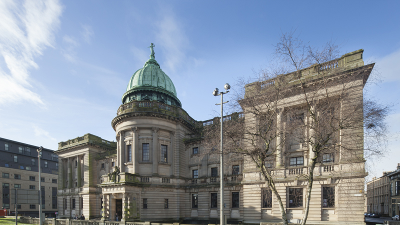 The Mitchell Library, Glasgow, on a sunny day. A large light sandstone building, with many windows and statues on the architecture of the building. Grass and trees are in front of the building.