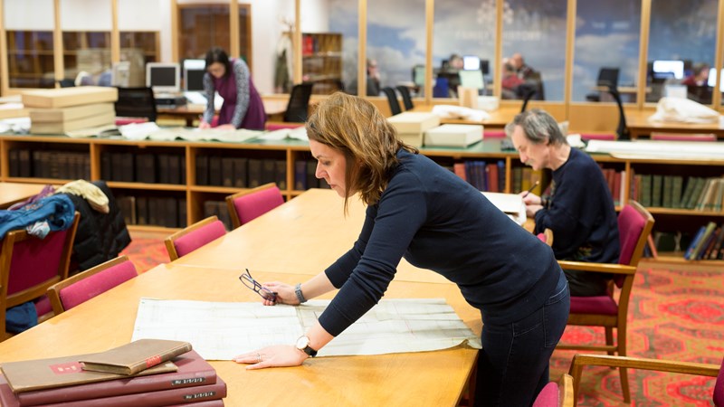 Woman in the Mitchell Library standing up with her glasses in her hand while looking at papers laid out on a desk 