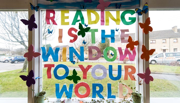 Multicoloured letters on a window in Barmulloch library - spelling out 'Reading is the window to your world' there are also coloured paper butterflies. You can see out in to the street beyond the library.