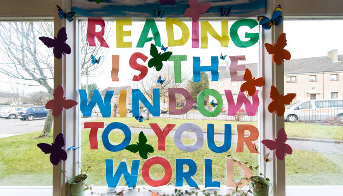 Multicoloured letters on a window in Barmulloch library - spelling out 'Reading is the window to your world' there are also coloured paper butterflies. You can see out in to the street beyond the library.