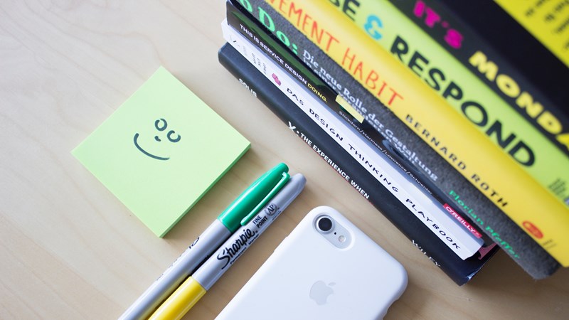 A pile of books sit next to a post it note, with a smiley face drawn on