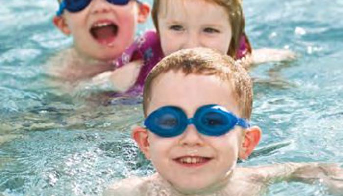 Three children in a swimming pool, two are wearing swimming goggles and all are smiling at the camera