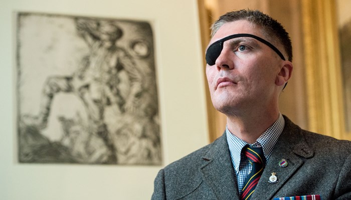 A man wears a suit, adorned with medals. He has an eye patch. He stands in front of a painting of a soldier in World War 1