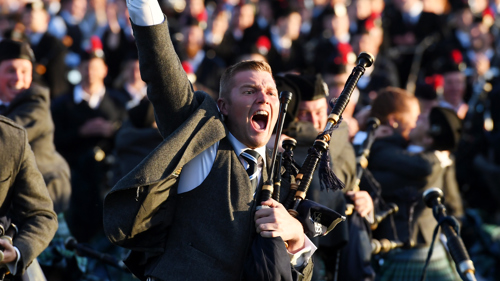 Piper wearing a kilt celebrating outdoors at the World Pipe Band Championships with lots of pipers all wearing the same tartan in the background