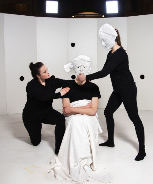 Three performers. They all wear black. Two wear masks, and one has a white sheet covering their legs.