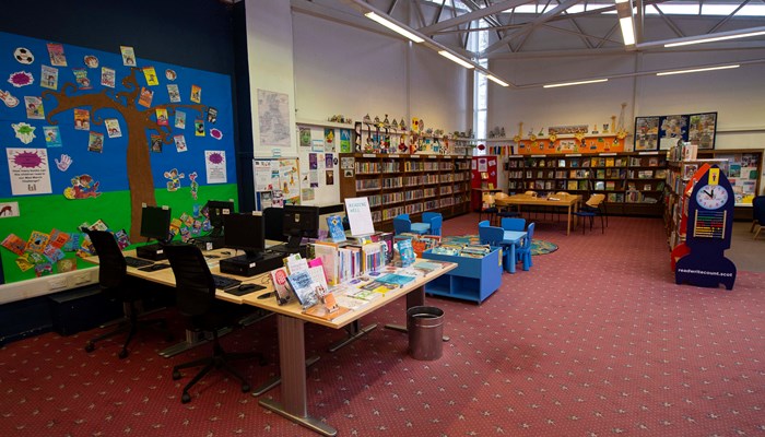 Main view of the reading and PC area of the library which has high ceilings and metal beams, a pink carpet and children's artwork on the wall