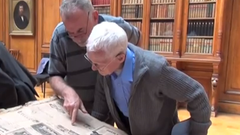Two men looking at a large publication containing historical information and black and white photos with bookcases of information behind them