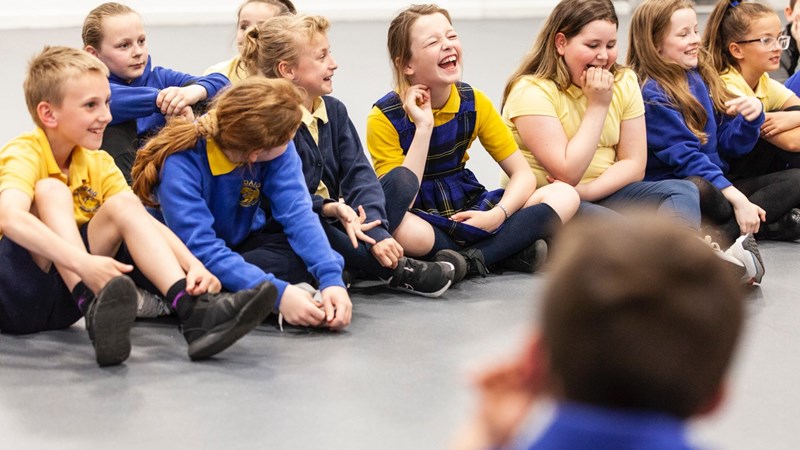 A group of school children sit together, laughing, in a school hall