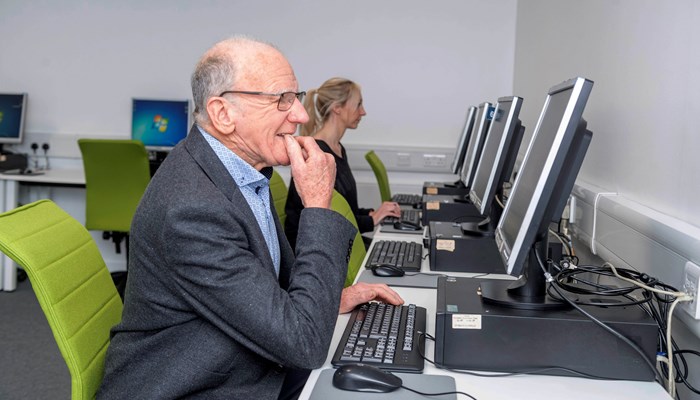 An older person wearing a grey suit looking at a computer screen with their hands on the keyboard in the library