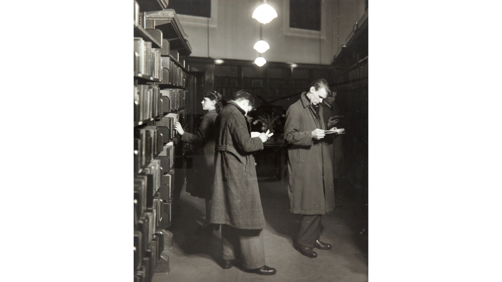 Black and white photograph of three patrons wearing heavy winter coats browsing books along tall wooden bookshelves inside a library aisle.