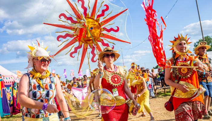 A vibrant parade moves across a grassy field under a sunny sky, featuring people in sun-themed costumes and a large, ornate sun prop held aloft.