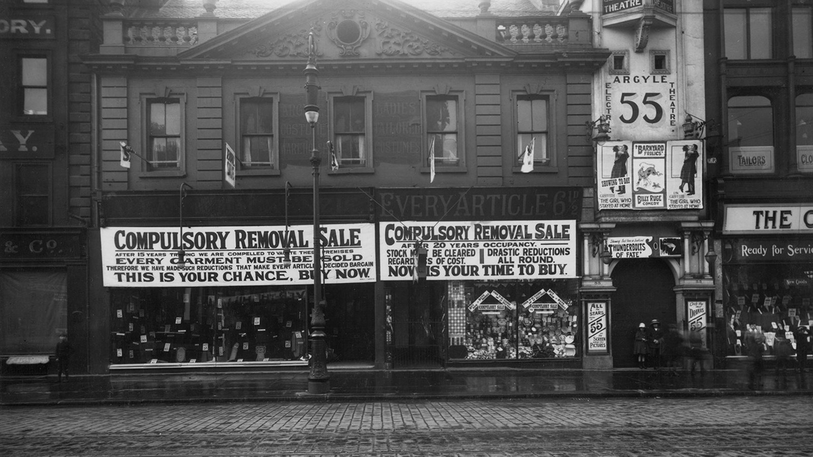 A black and white photograph of Argyle street which has a cobbled road showing a theatre entrance. Other shops have lots of advertisements in their windows.