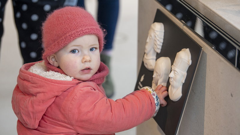 A young child wearing a pink jacket and hat touching a piece of wall art of the wall whilst staring straight into the camera