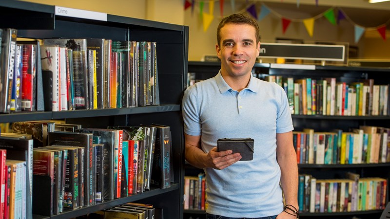 A person standing and leaning against a bookshelf in a library. They are holding an e-reader and smiling.