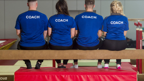 4 gymnastics coaches sitting on balance bar with their backs to us with 'Coach' on the back of their uniform