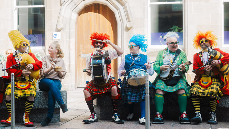 A street band wearing vibrant, oversized wigs, sunglasses, and traditional kilts plays instruments—including bagpipes, drums, and a banjo—while seated on an outdoor stone bench next to a spectator.