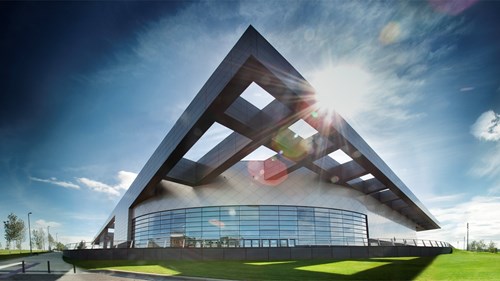 panoramic shot of the exterior of emirates arena. the building is grey and is in the shape of a triangle. there is green grass surrounding the building