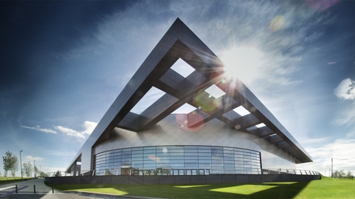 panoramic shot of the exterior of emirates arena. the building is grey and is in the shape of a triangle. there is green grass surrounding the building