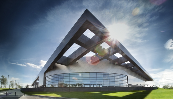 panoramic shot of the exterior of emirates arena. the building is grey and is in the shape of a triangle. there is green grass surrounding the building