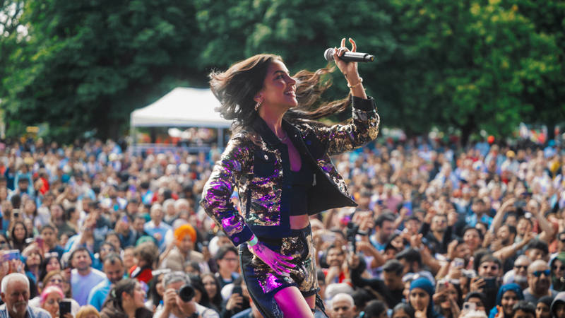A singer in a sequined jacket holds a microphone and performs to a large outdoor crowd gathered in a park setting.