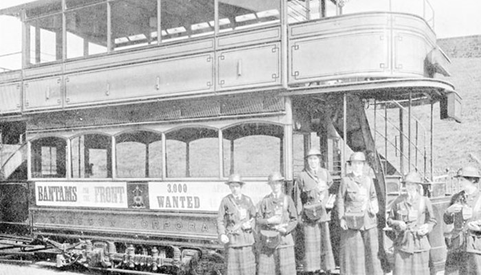 A black and white stationary tram with advertisement on the side, with 6 women standing at the doorway posing for a photo.