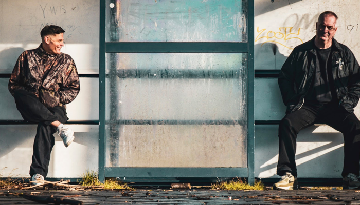 Two men sit on benches inside a weathered glass bus shelter, one on the left smiling in a camouflage jacket with legs crossed, and the other on the right looking forward in a dark jacket.