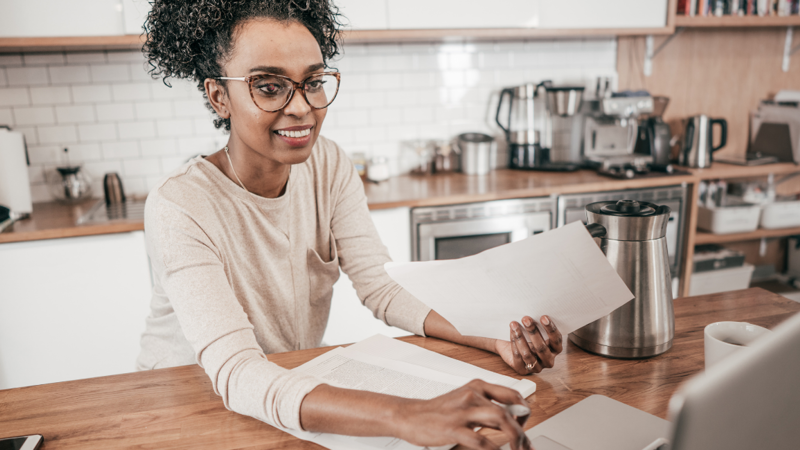 A person is working on a laptop from their kitchen. They have papers in their hand and smiling towards the screen. 