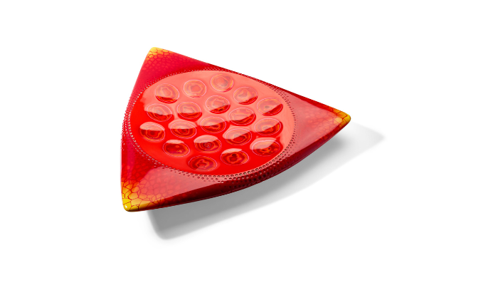 Triangular red glass dish with softly curved sides and rounded corners, shown against a light grey background. The translucent surface features a cluster of raised circular forms in the centre, surrounded by a dotted border. Gradients of red, orange and yellow create a glowing effect, while subtle crackle-like patterns appear near the edges.