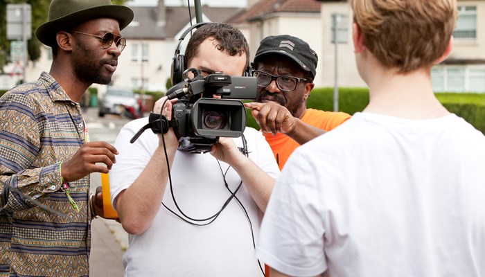 Person holding a video camera with two people on either side giving advice. The back of the person being filmed is also in view.