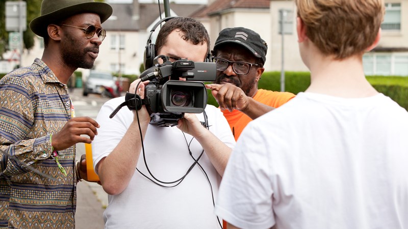 Person holding a video camera with two people on either side giving advice. The back of the person being filmed is also in view.