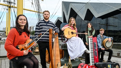 A group of five musicians gathered on the deck of a tall ship in front of a Riverside Museum in Glasgow. They are holding harps, guitars, violins, keyboard and drum.