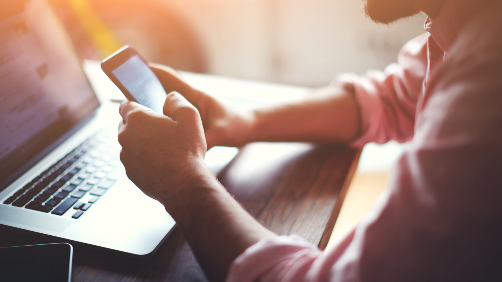 stock photograph of a person using a mobile phone while a laptop sits behind it.