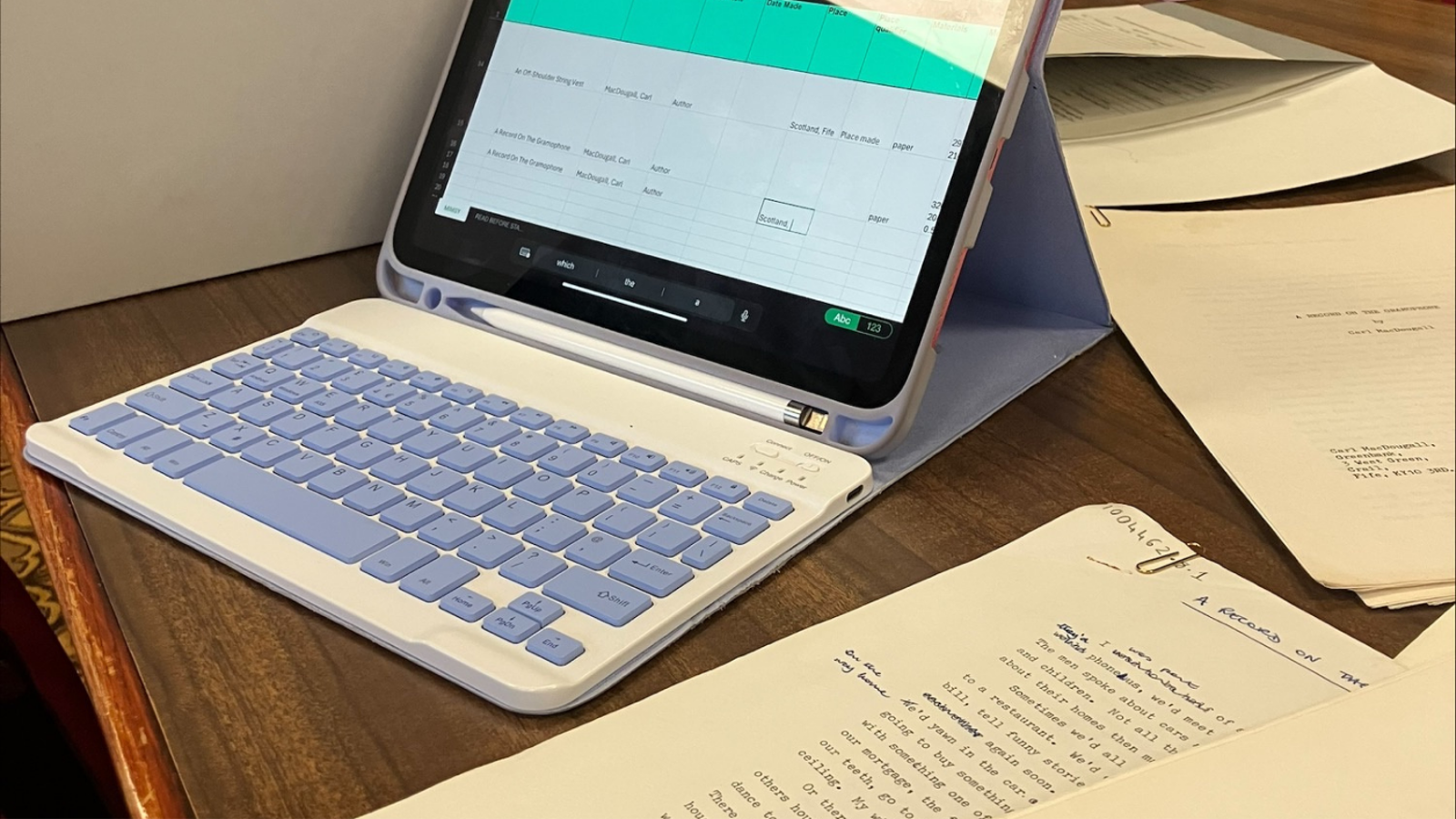 A tablet in a light blue case displays a spreadsheet listing multiple entries for “A Record on the Promises” by Carl MacDougall. The tablet sits on a wooden table surrounded by printed manuscript pages of the same title, some with typed text and handwritten annotations.