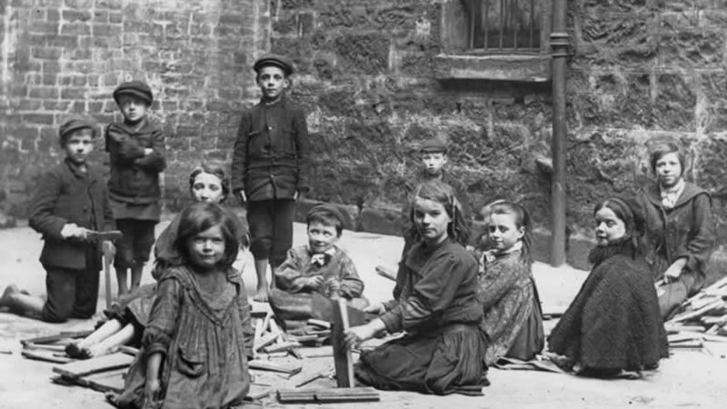 A black and white photo of children sitting in a courtyard of a large building with no shoes on breaking sticks.