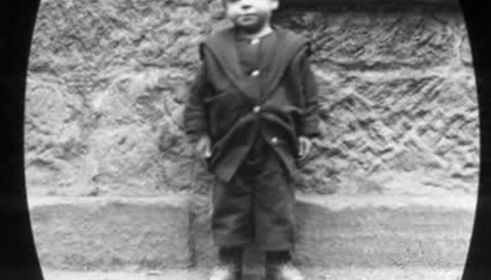 A black and white photo of a young child posing against an exterior house wall.