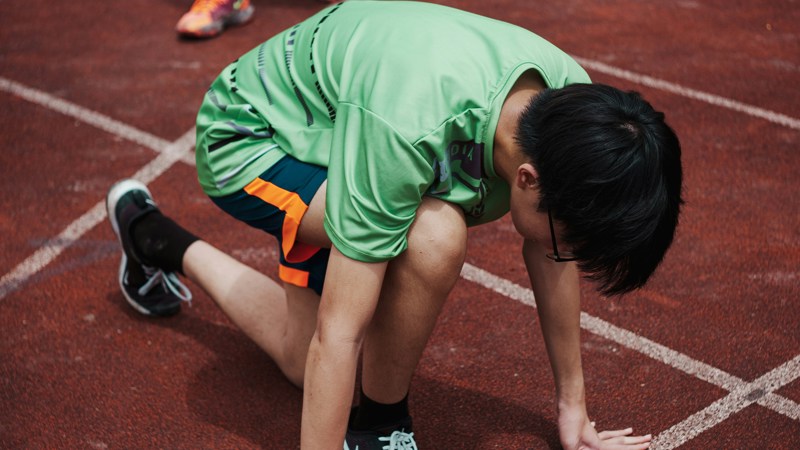 Participant in a green sports shirt crouches in a starting position on a red running track, with other runners in green in the background.