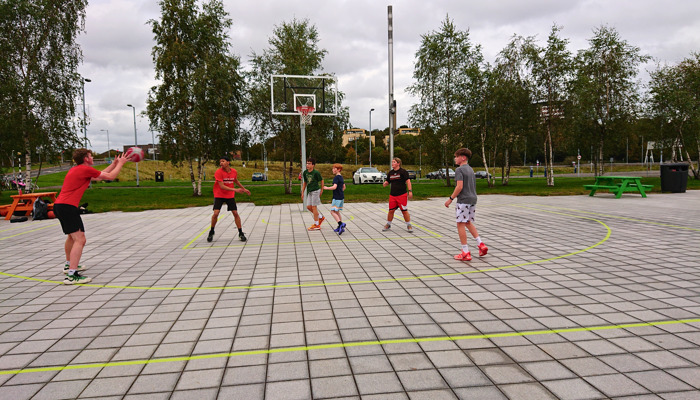 Five young people play basketball on an outdoor court with trees and buildings in the background.