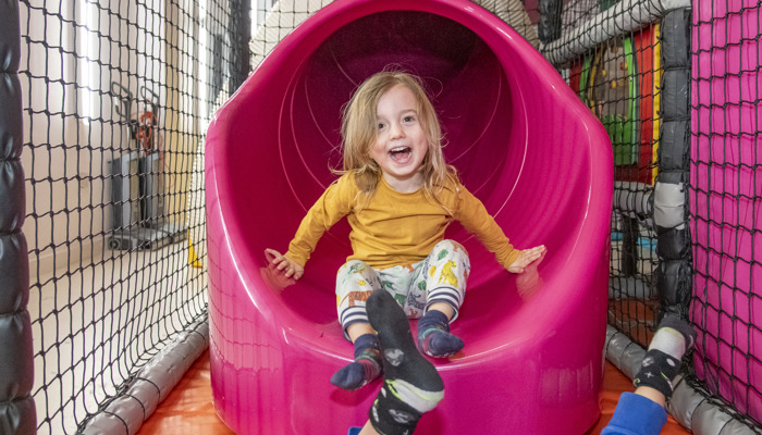Child exiting a pink tube slide in an indoor soft play centre, with netted walls and play structures visible around the slide.