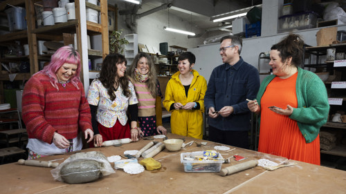 A group of six artists standing around a large wooden table in a studio space. They are smiling and working with clay