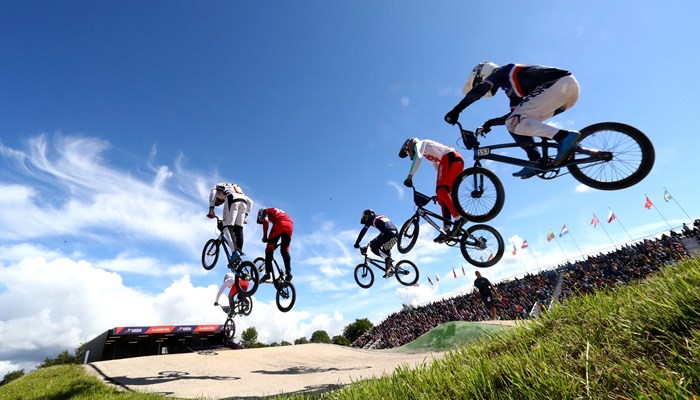 5 people on bikes going over jumps at a track. they are outdoors and are wearing safety helmets