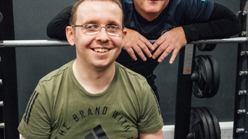 two men looking at the camera and smiling in a gym setting, one man is standing and the other is in a wheelchair, the main in the wheelchair has had his arm amputated from below the elbow. 