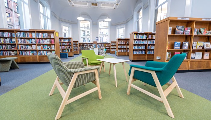 Internal photo of a library with large wooden bookshelves filled with colourful books, large windows and cosy green toned armchairs surrounding a coffee table on a green floor runner.