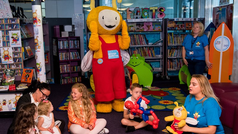 Adults and children sitting around in a circle within a library setting holding toys and books with the Book bug mascot standing behind them.